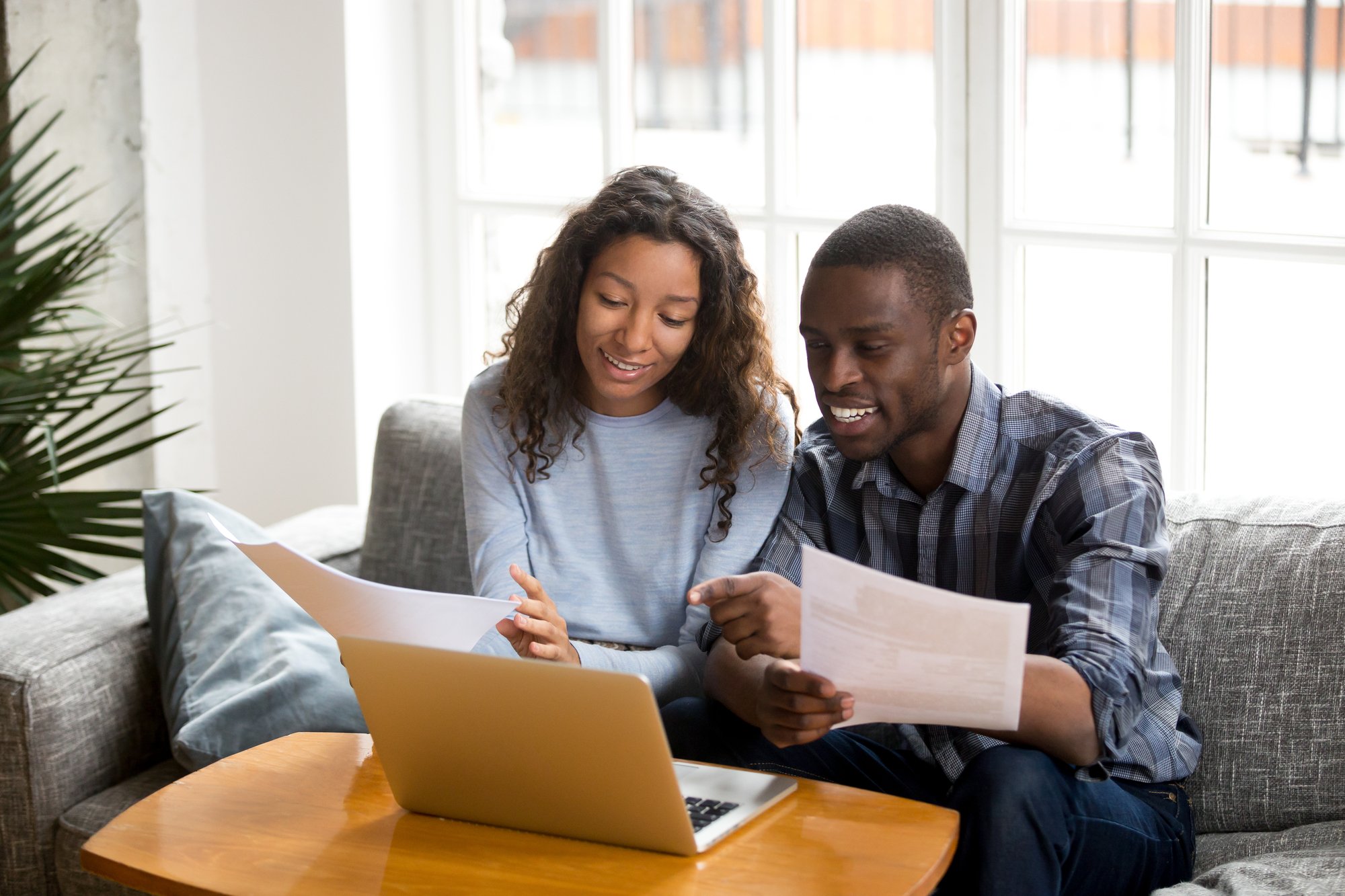 African black positive married couple sitting on sofa at home read documents paper checking bills, bank account balance feeling satisfied and happy