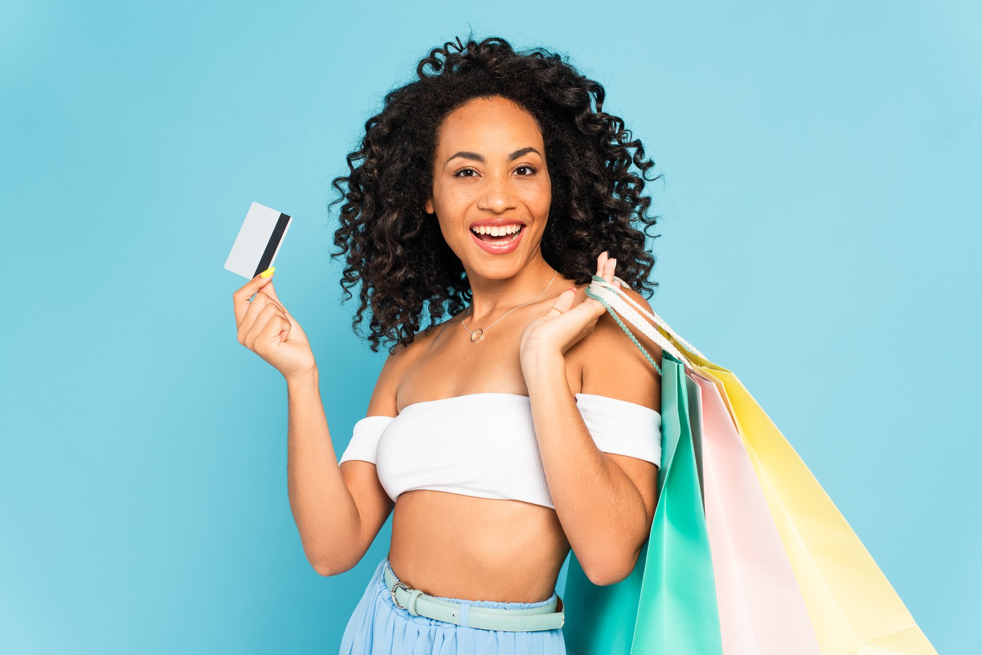 Excited african american woman holding credit card and shopping bags isolated on blue