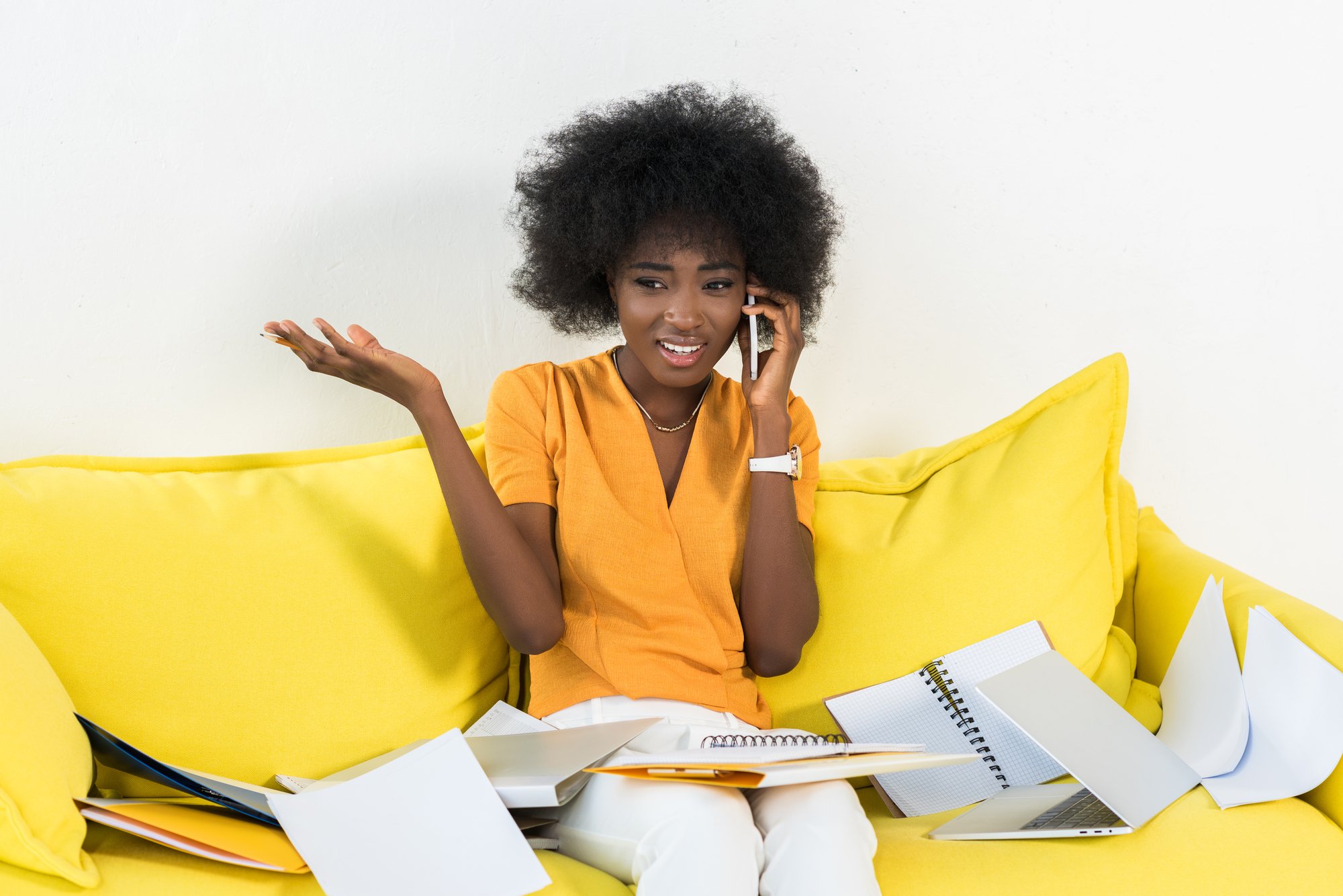 Portrait of confused african american freelancer talking on smartphone on sofa with papers at home