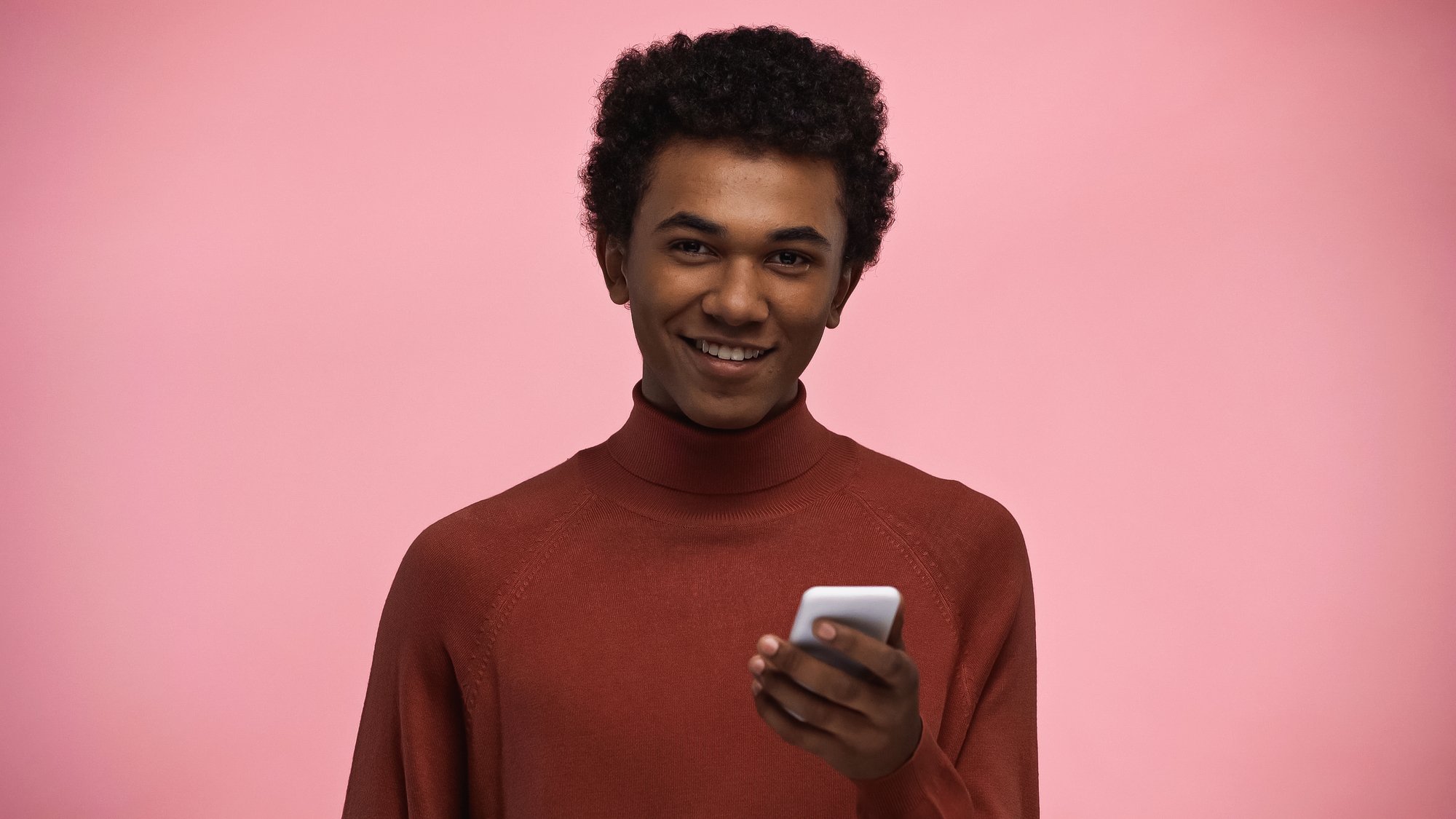 Smiling african american teenager in turtleneck sweater and glasses looking at camera while holding smartphone isolated on pink