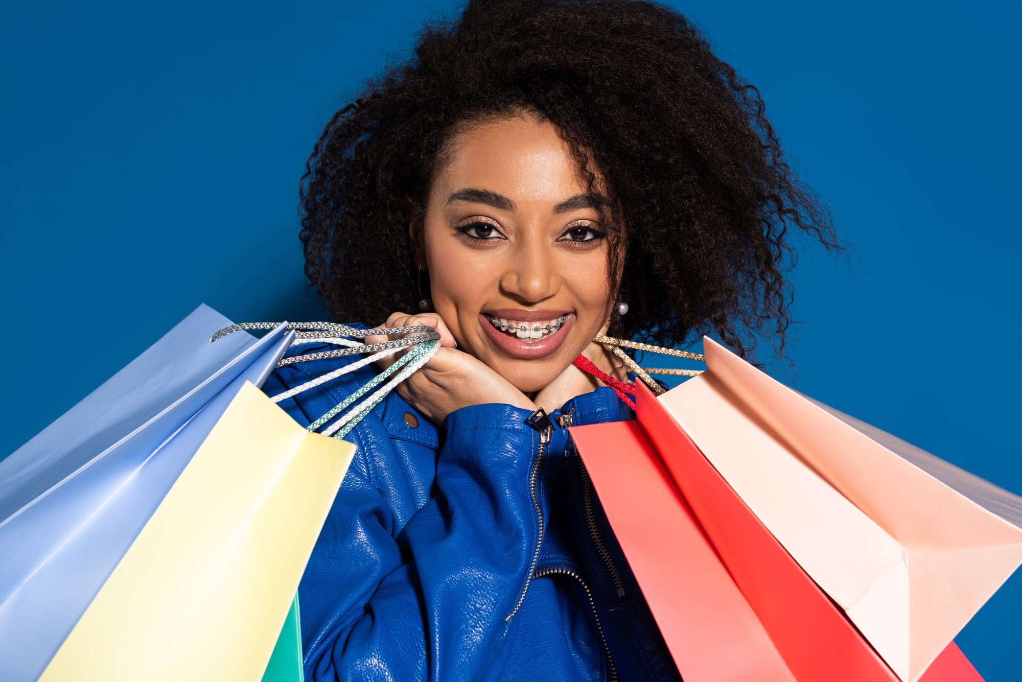 Smiling african american woman with braces and shopping bags isolated on blue
