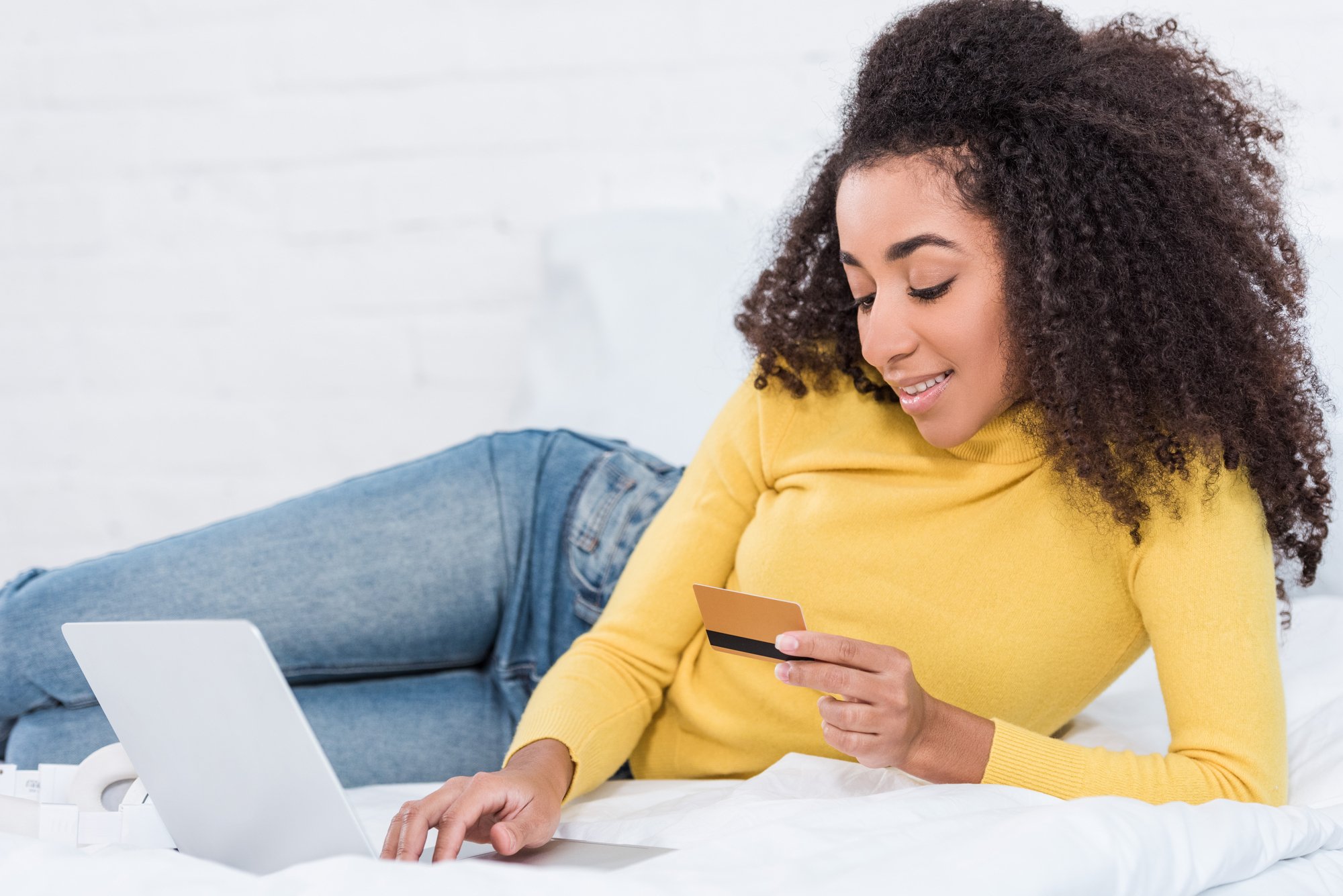 Young attractive african american woman holding credit card and doing online shopping on laptop in bed at home