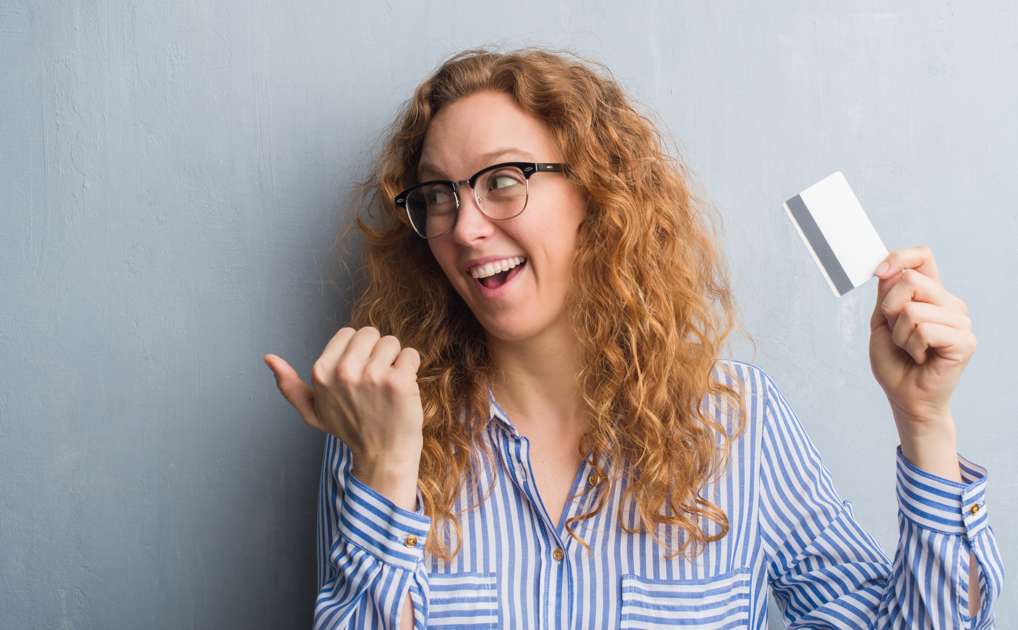 Young redhead woman over grey grunge wall holding credit card pointing and showing with thumb up to the side with happy face smiling